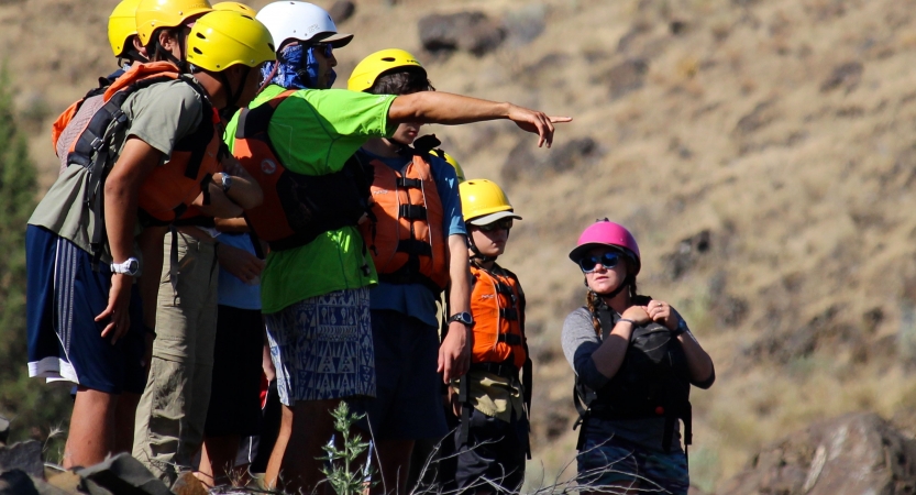 A group of students wearing life jackets and helmets stand in a line, possibly above a river. One of them is pointing at something in the distance.